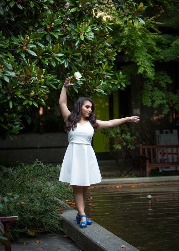 Raya Tuffaha in a white dress and blue shoes, posing in North Seattle, in front of greenery, in a ballet position