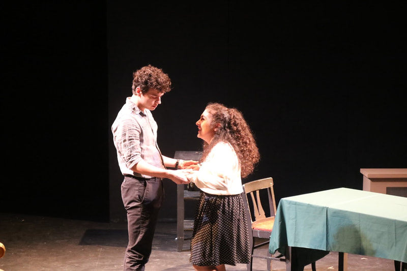 Raya (Catherine) smiling with Timothy St. Pierre (Eddie), wearing a white shirt and black skirt. Timothy holds Raya's hands.