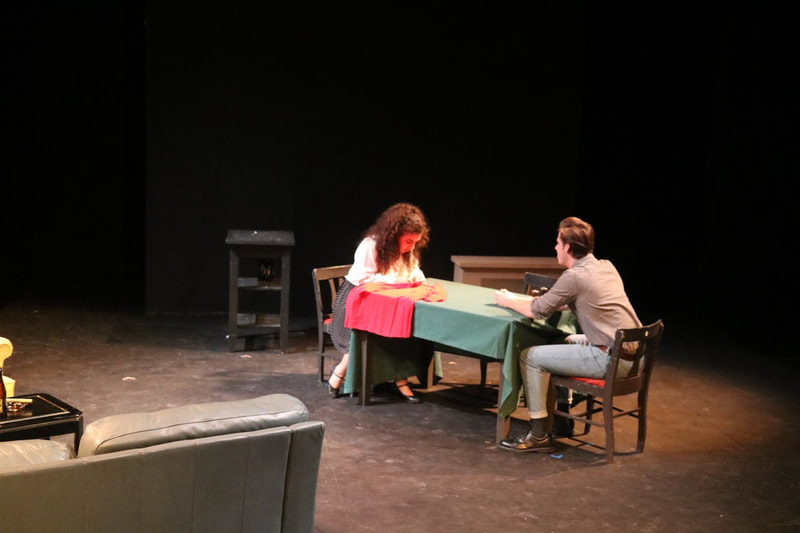 Raya Tuffaha sitting at a table with a green tablecloth, sewing a red shirt, she is wearing a white shirt and black skirt, sitting opposite her scene partner who is wearing a gray shirt and jeans