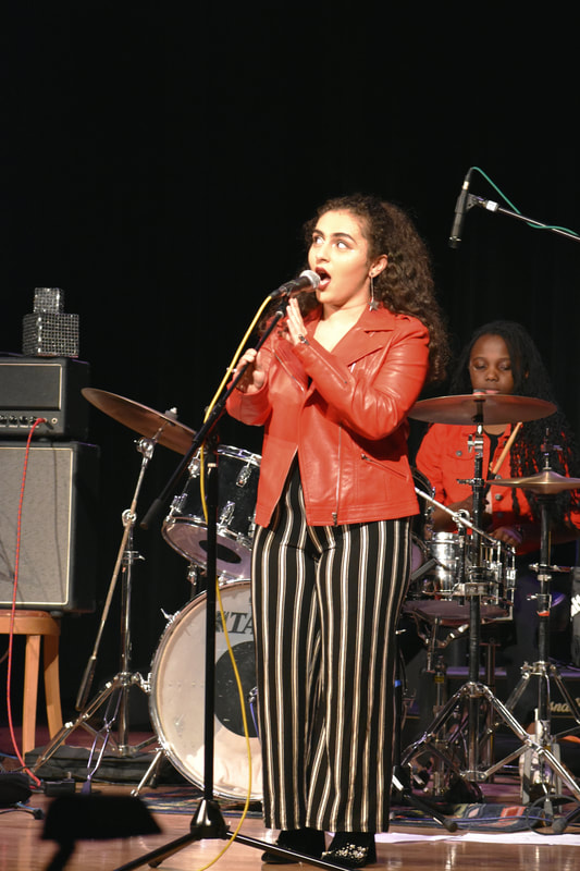 Raya Tuffaha at a microphone, wearing a red leather jacket, black and white striped jumpsuit, she is singing. Drummer in the back is wearing a red jacket and has braids in her hair