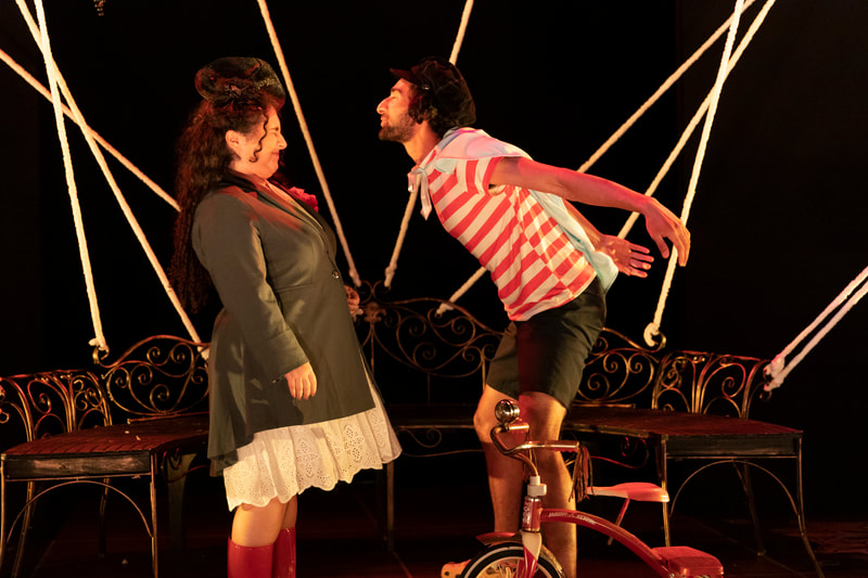 Raya (Eurydice) and Nikhil Tignor (Child) face each other. Nikhil, wearing a red and white striped shirt and blue cape, blows air in Raya's face the way someone blows out birthday candles. Raya wears a gray jacket, white skirt and black fascinator hat. Both people's eyes are closed.