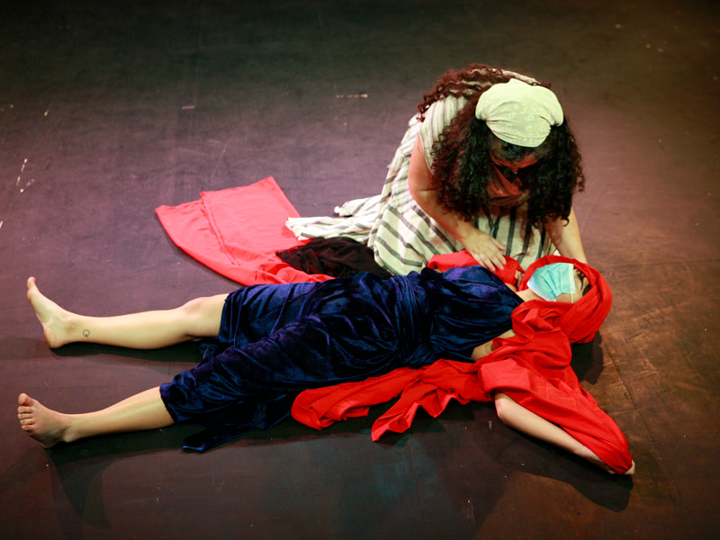 Raya (Nurse), wearing a striped skirt and a bandana, kneels over Nena Martin (Phaedra), wearing a dark blue dress and lying on the ground. Nena is wrapped in a red fabric.