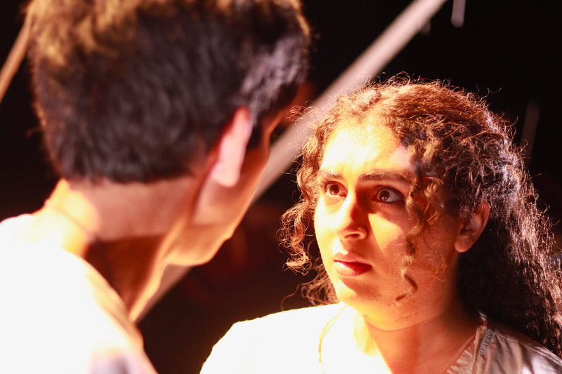 Raya (Eurydice), looking at Daniel Oakes (Orpheus) with fear and regret. The lighting is warm and golden. Both people are wearing white. Only Raya's face is visible.