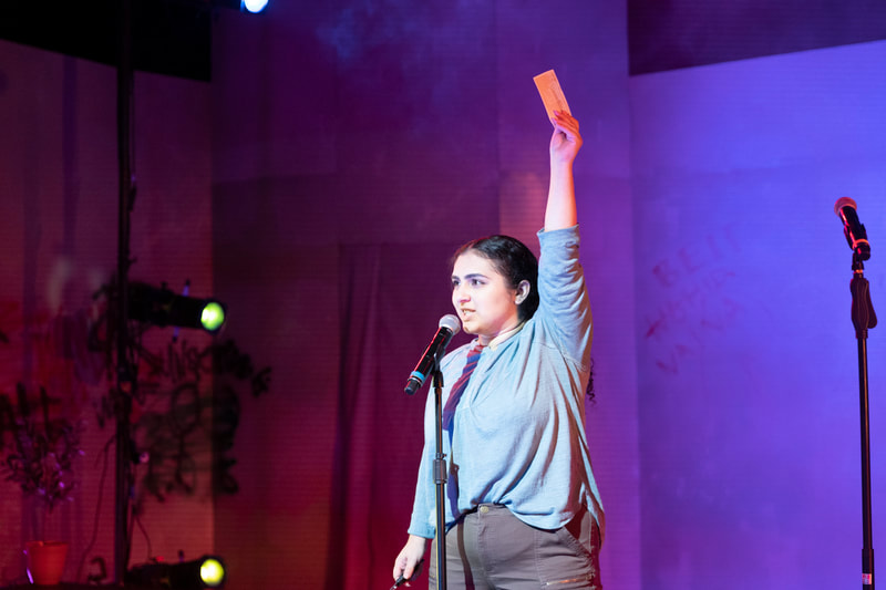 Raya (Naina) stands at a microphone, wearing a blue-green shirt. Her left arm is raised directly above her head. She holds a photo in her left hand.